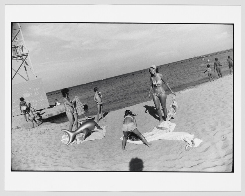 Black and white photo of beachgoers on sandy shore with a lifeguard tower to the left, inflatable dolphin toy on the ground, and calm sea in the background. People are seen sunbathing, walking, and engaging in beach activities on a sunny day.
