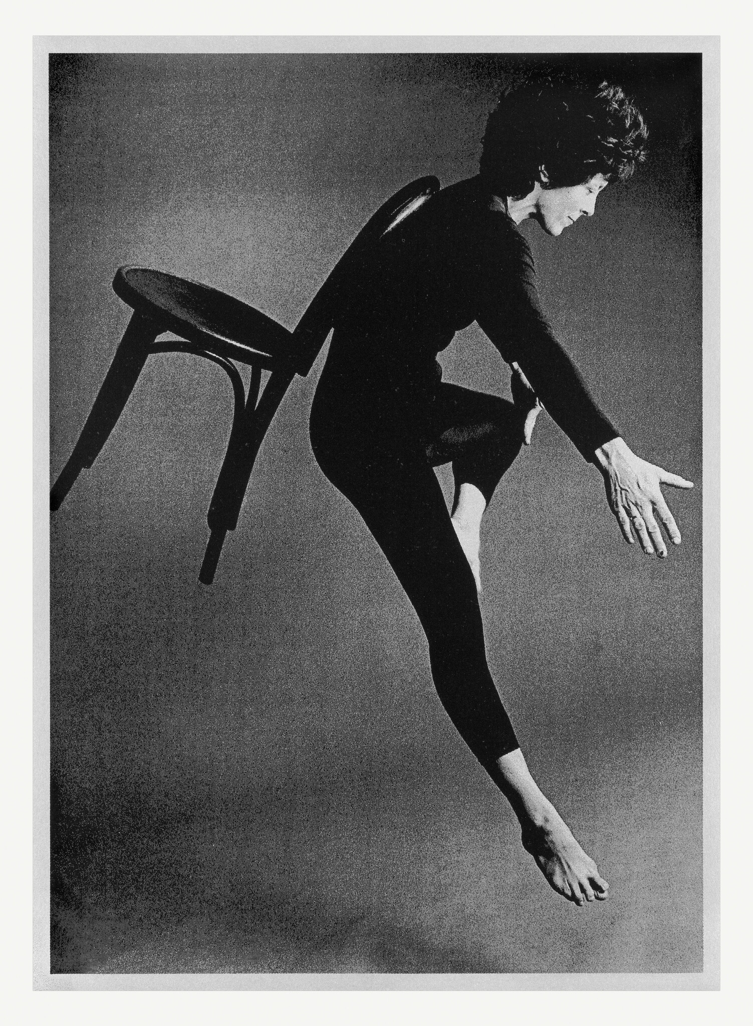 Woman in black leotard suspended midair with a chair hooked over her back, reaching forward.