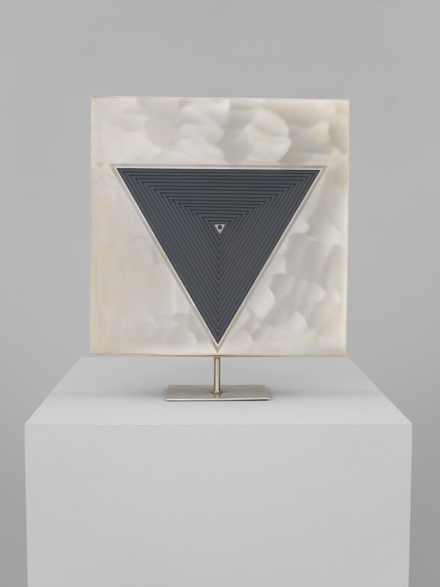 A square metallic panel on a stand features a large inverted triangle of concentric lines.