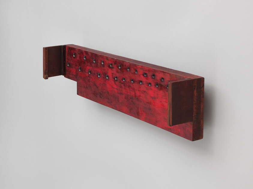 A long red wooden wall shelf with metal studs and dark end brackets mounted on a white wall.