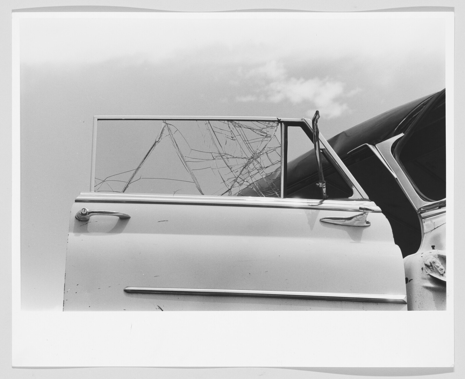 A damaged car door with a cracked window and bent frame against a cloudy sky.