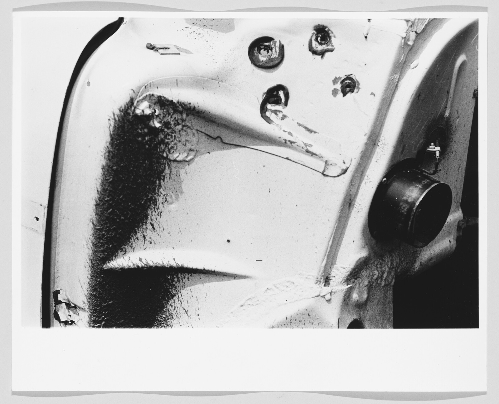 Dented white metal panel with burn marks, peeling paint, and a round black cap attached.