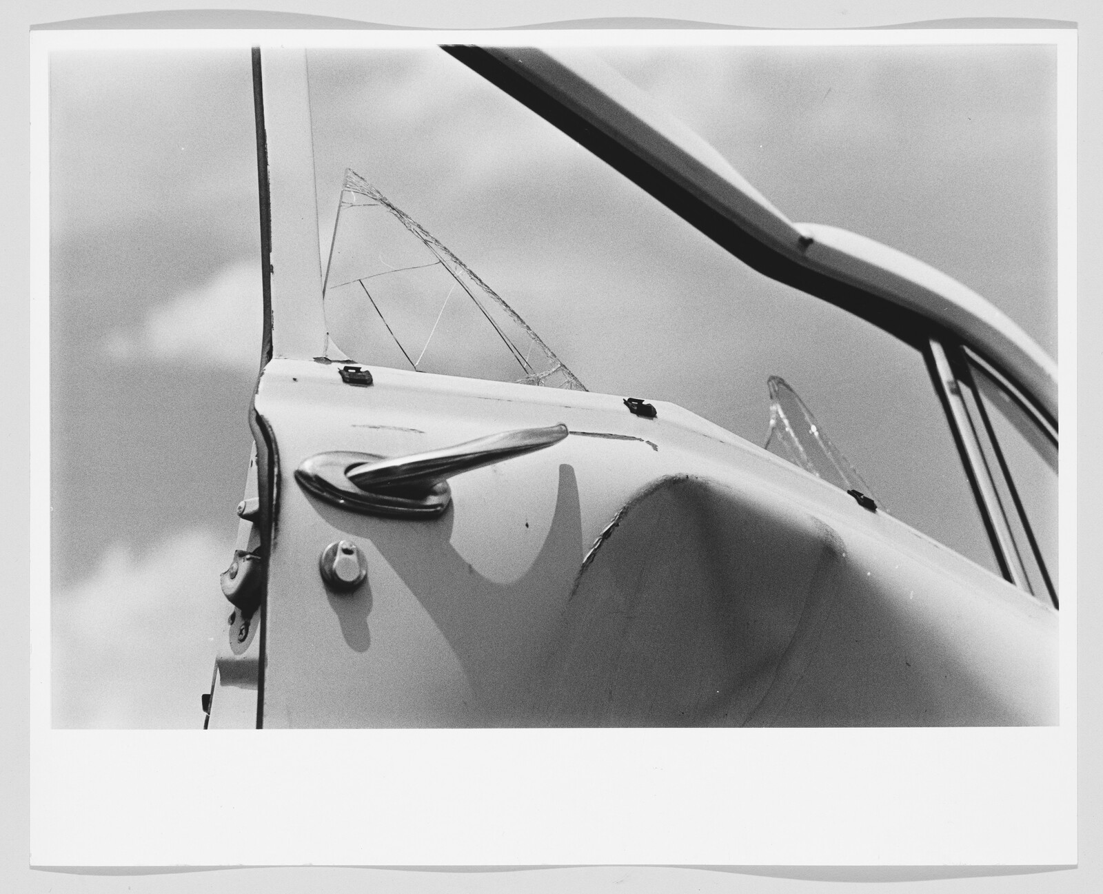 Car door with a deep dent and two shattered vent windows under a cloudy sky.