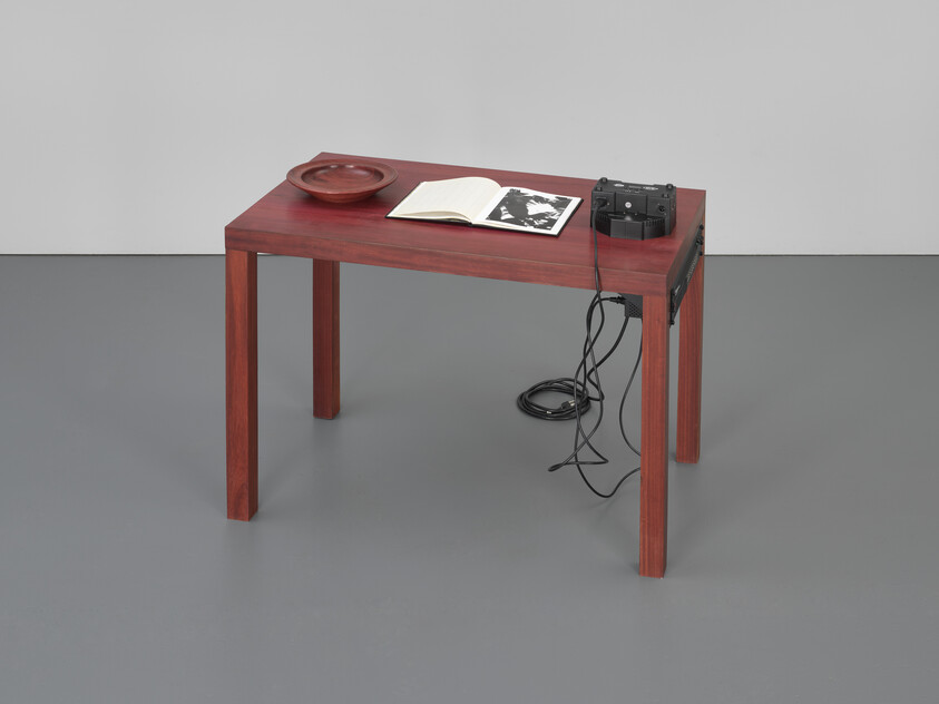 A simple wooden table holds an open book, a shallow bowl, and a vintage tape recorder with tangled cords.