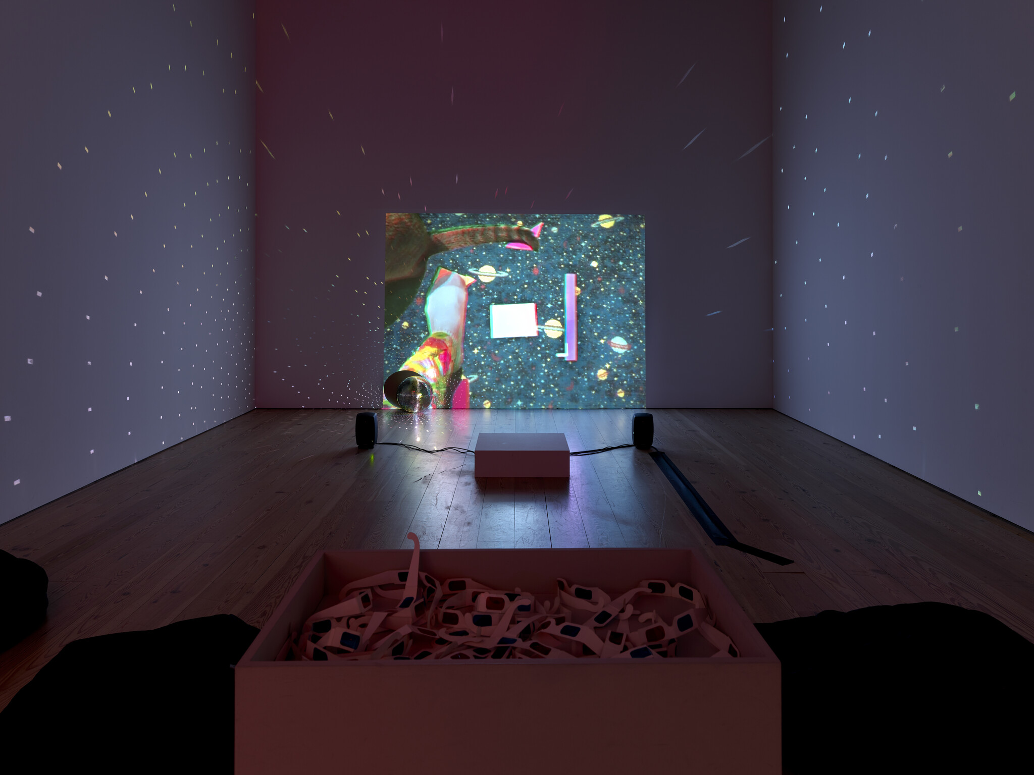 A dark gallery room with a projector displaying a colorful space scene and a box of 3D glasses.