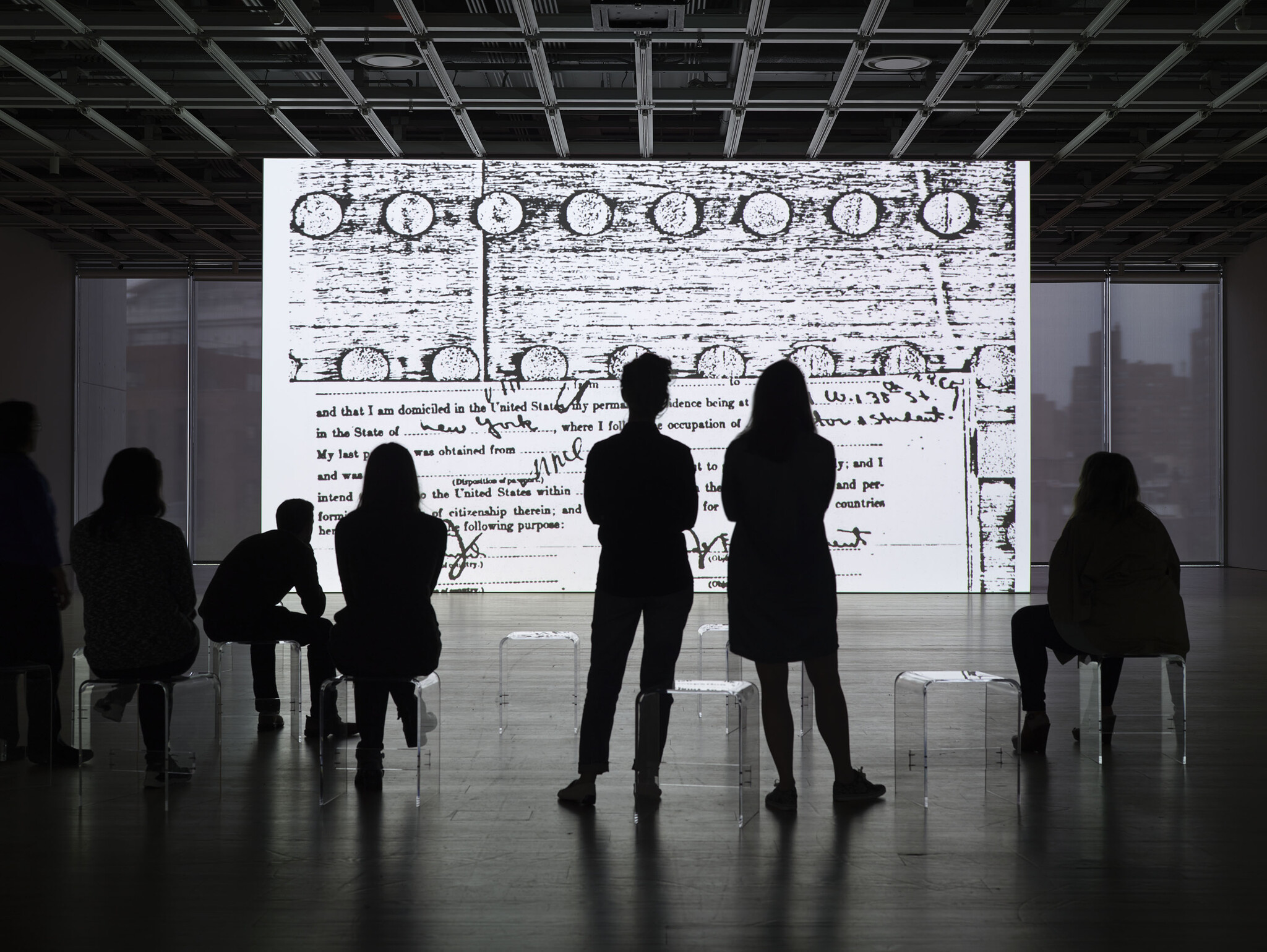 Silhouetted figures of people sitting and standing in an art gallery, observing a large, illuminated wall projection of a document with text and circular stamps. The room is dimly lit, emphasizing the contrast between the bright projection and the dark outlines of the viewers.