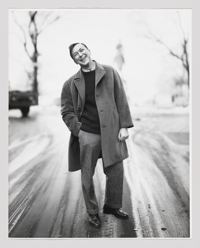 A short-haired white man in a peacoat, sweater, slacks, and oxfords stands on an unpopulated road with one hand in his pocket, his head titled as he smiles brightly.