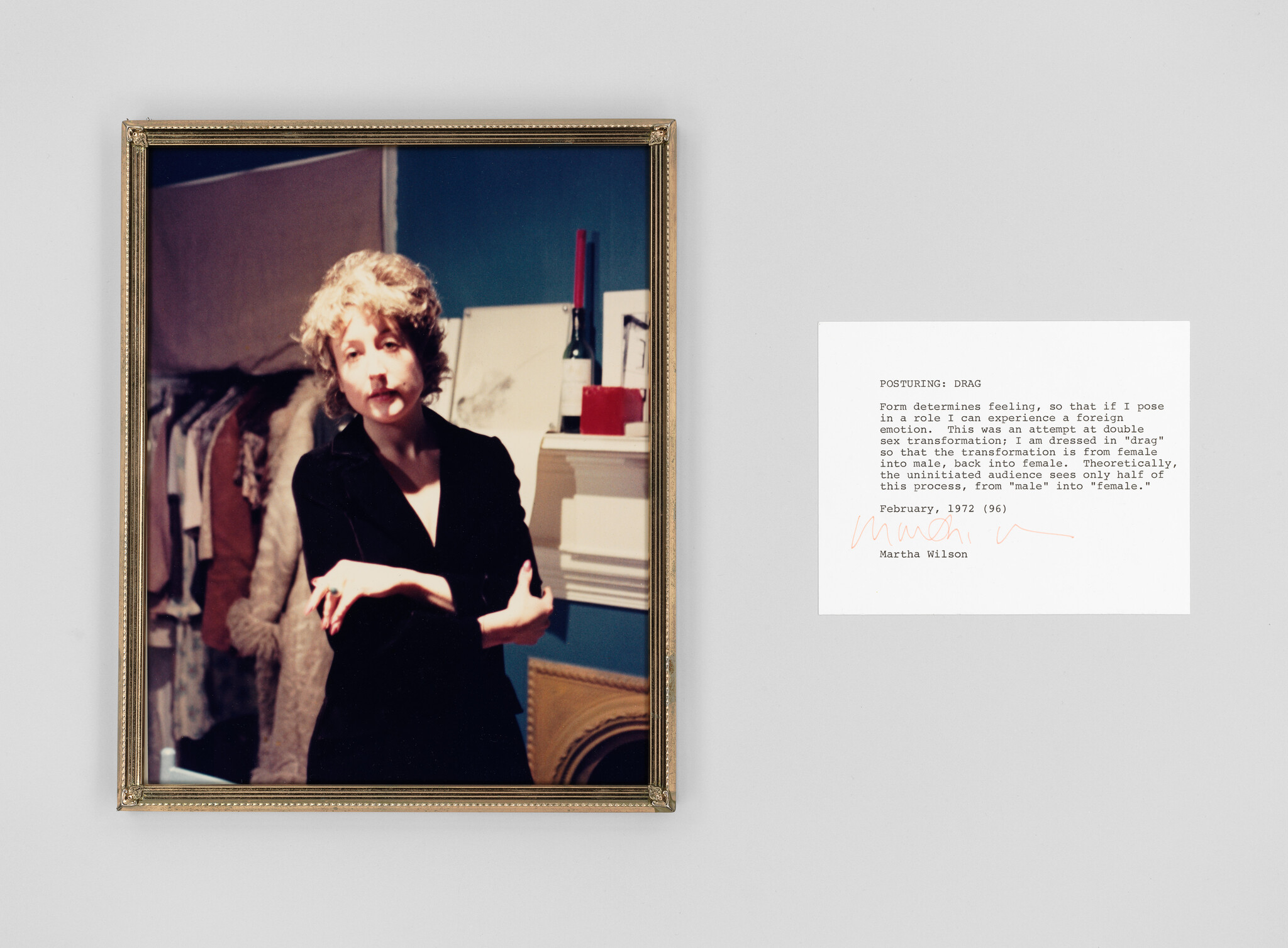 A framed photograph on a wall depicting a person with a mustache and short curly hair, dressed in a black blazer, posing with one arm crossed over their chest and the other resting on it. To the right of the photograph is a white card with typewritten text and a signature, explaining the concept of "posturing" in drag to express the fluidity of gender roles.
