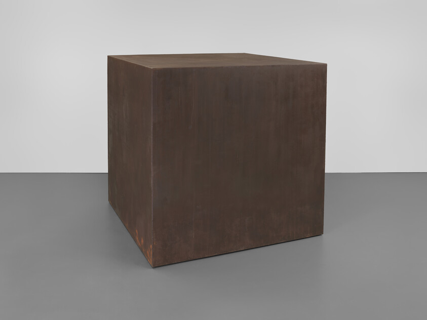 A large rust-colored metal cube sits on a gray floor against a white wall.