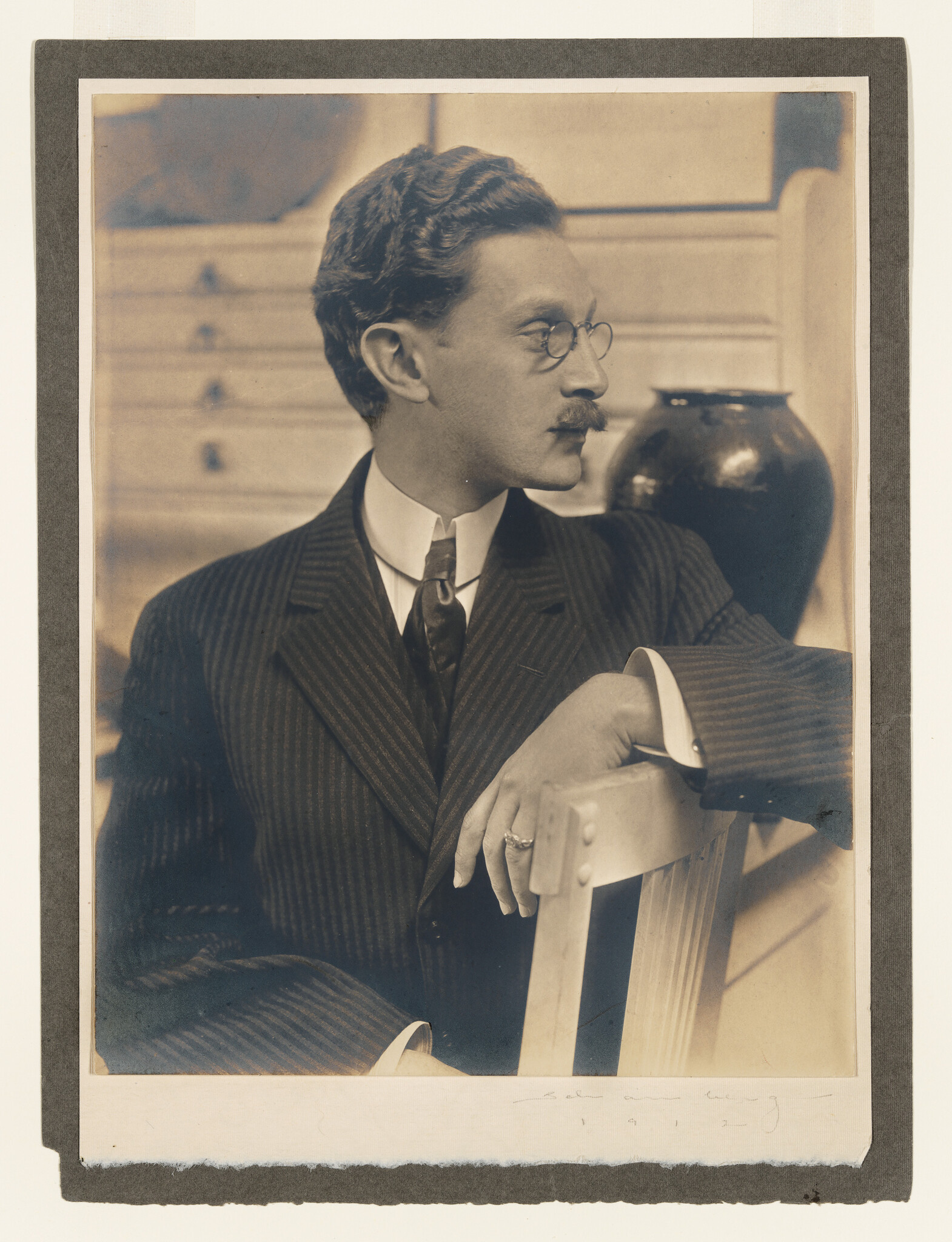 Man in pinstripe suit and round glasses sits with arm draped over chair, looking right.
