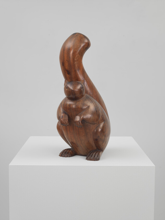 A carved wooden squirrel sculpture with a high curved tail sits on a white pedestal.