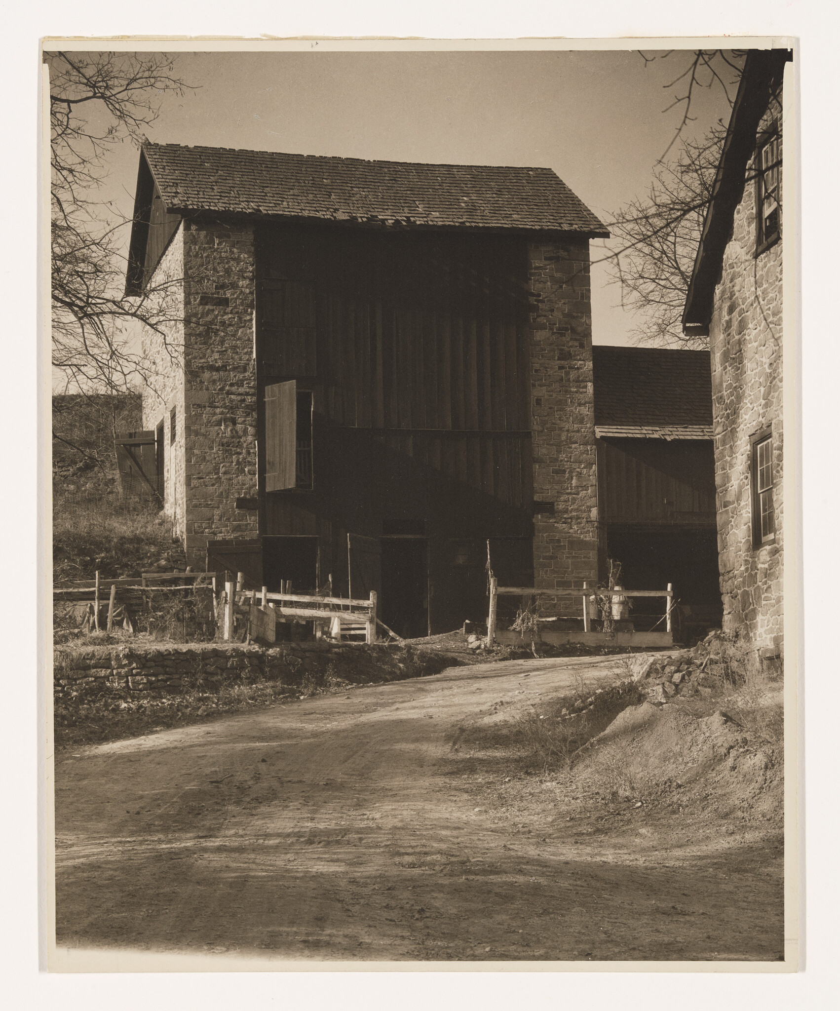 A dirt road leads up to a large stone barn with open wooden doors and fences.