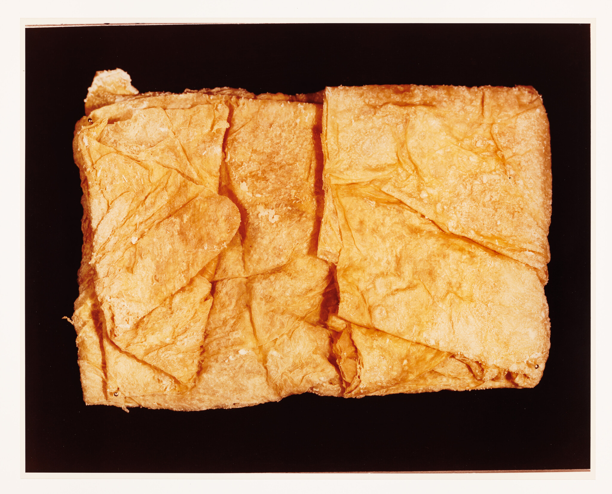 A close-up photograph of a large, square piece of flaky pastry with golden-brown coloring, displayed against a dark background. The pastry has visible layers and a textured surface with some crumbs.