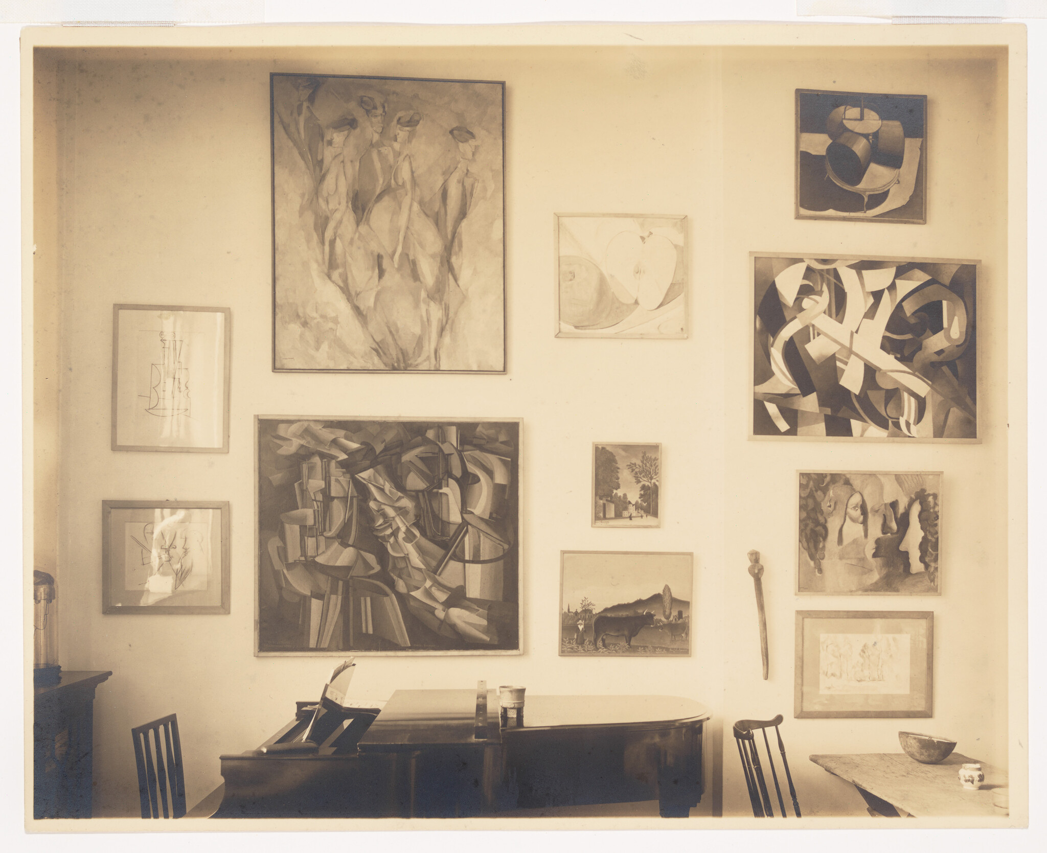 Grand piano and chairs beneath a wall displaying various framed abstract and figurative paintings.