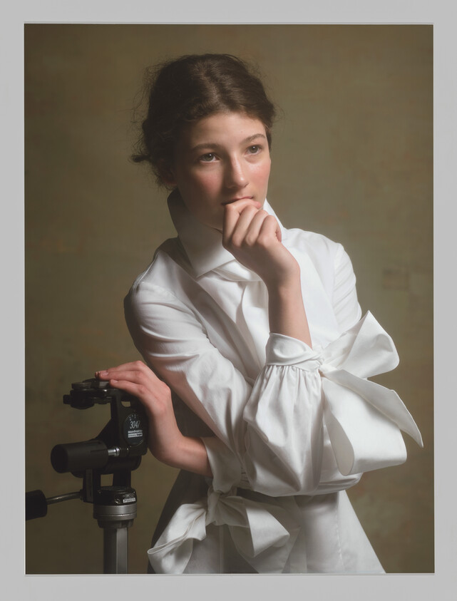 Young woman in a white blouse rests her hand on a tripod and looks thoughtful.