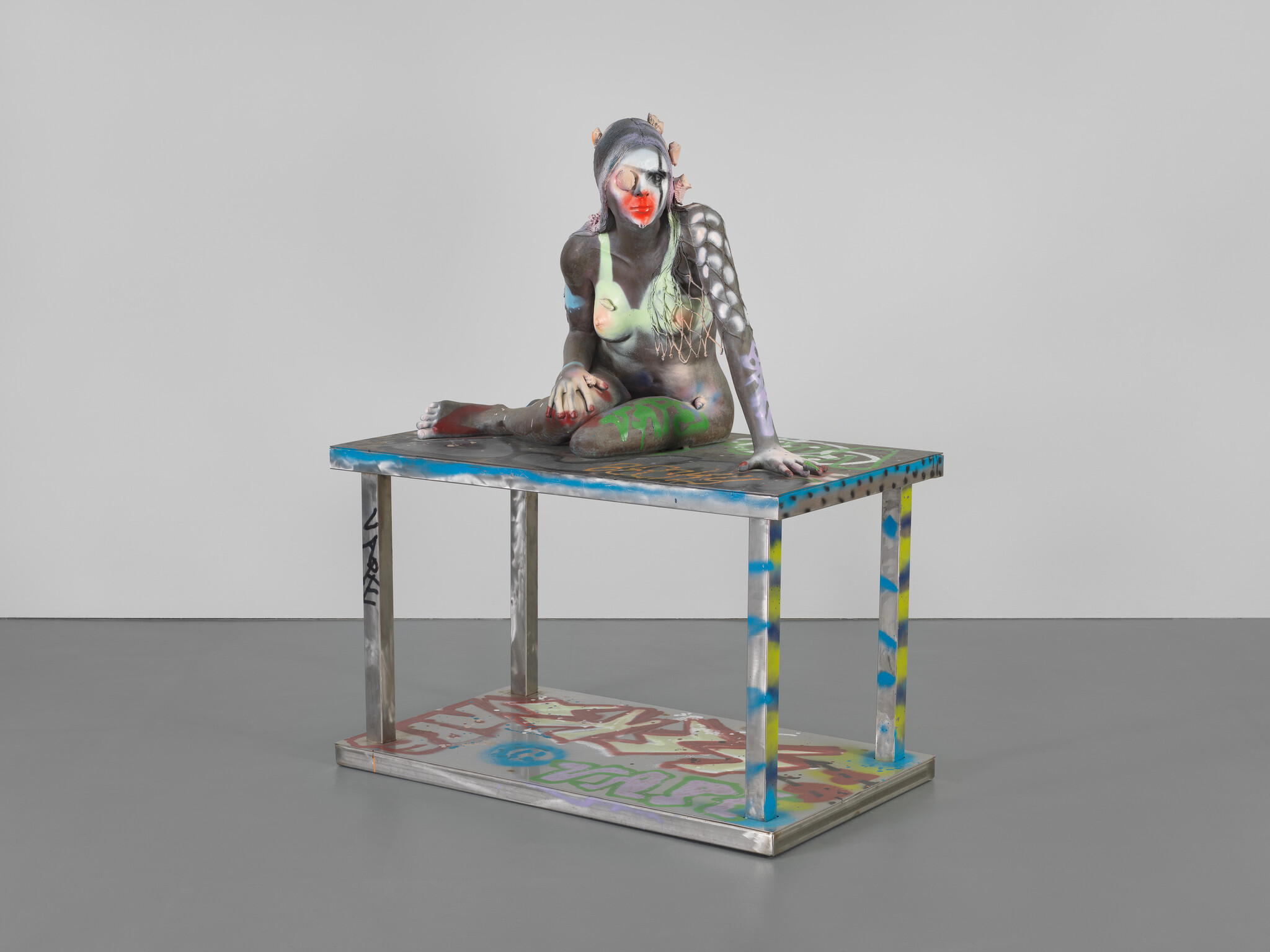 A painted female sculpture sits cross-legged on a graffiti-covered metal table in a gallery.
