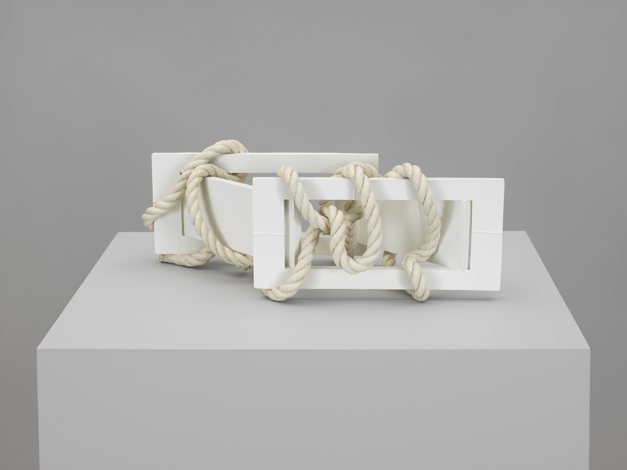 Two white rectangular wooden frames are intertwined and wrapped with thick cream rope on a pedestal.