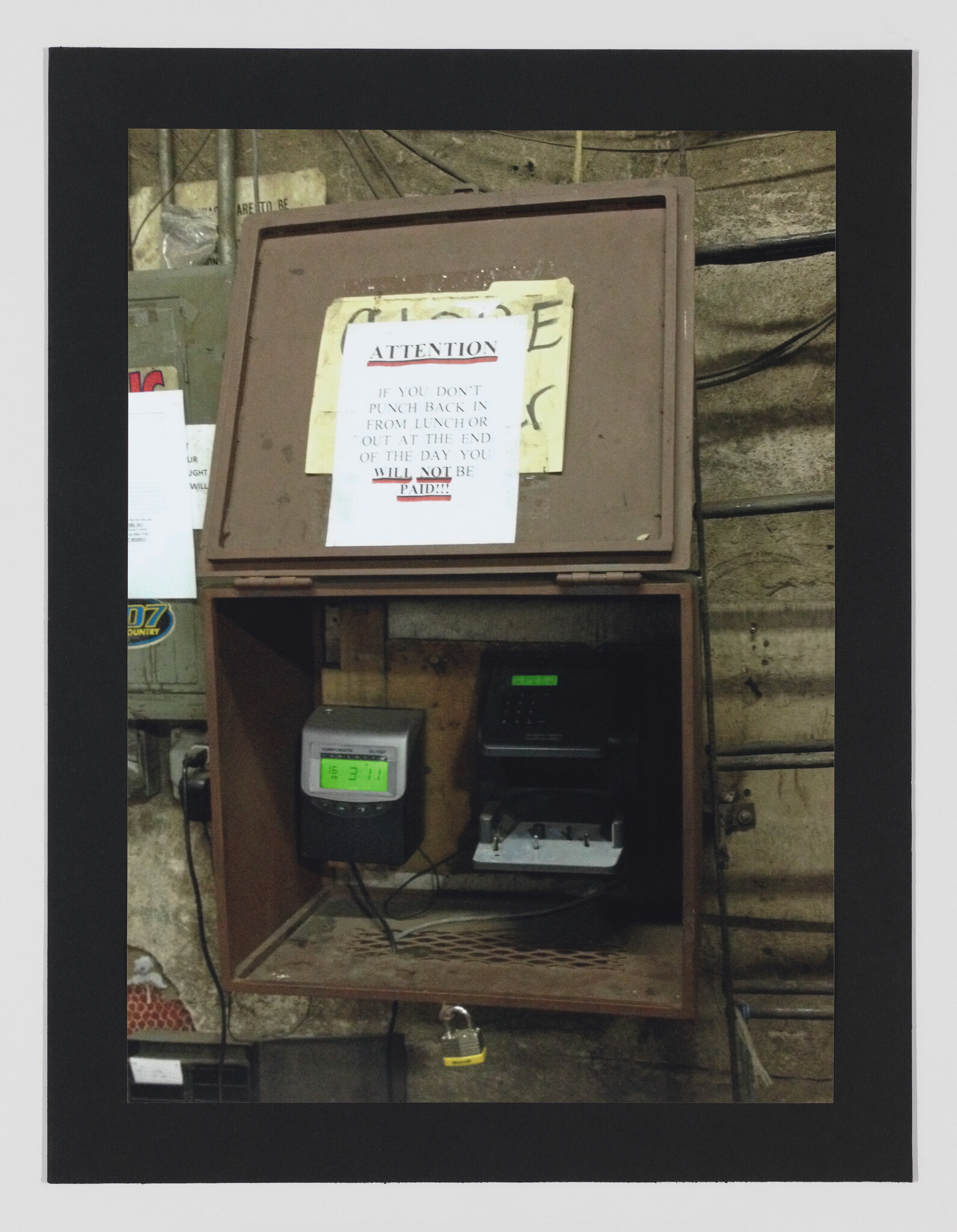 Wall-mounted time clock and punch machine inside a metal box with a warning sign about unpaid time.