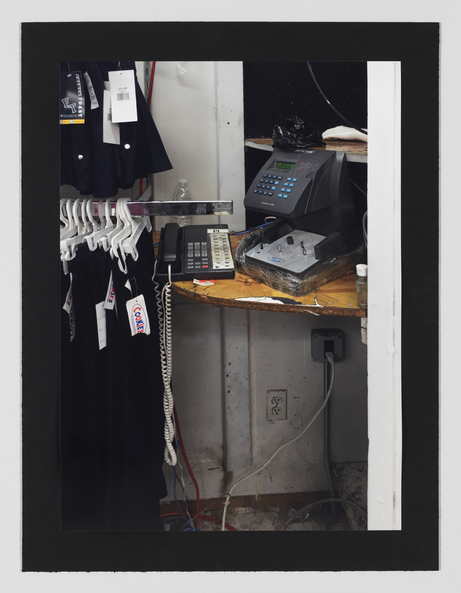 A cluttered back counter with a telephone, time clock machine, and hanging clothes on plastic hangers.