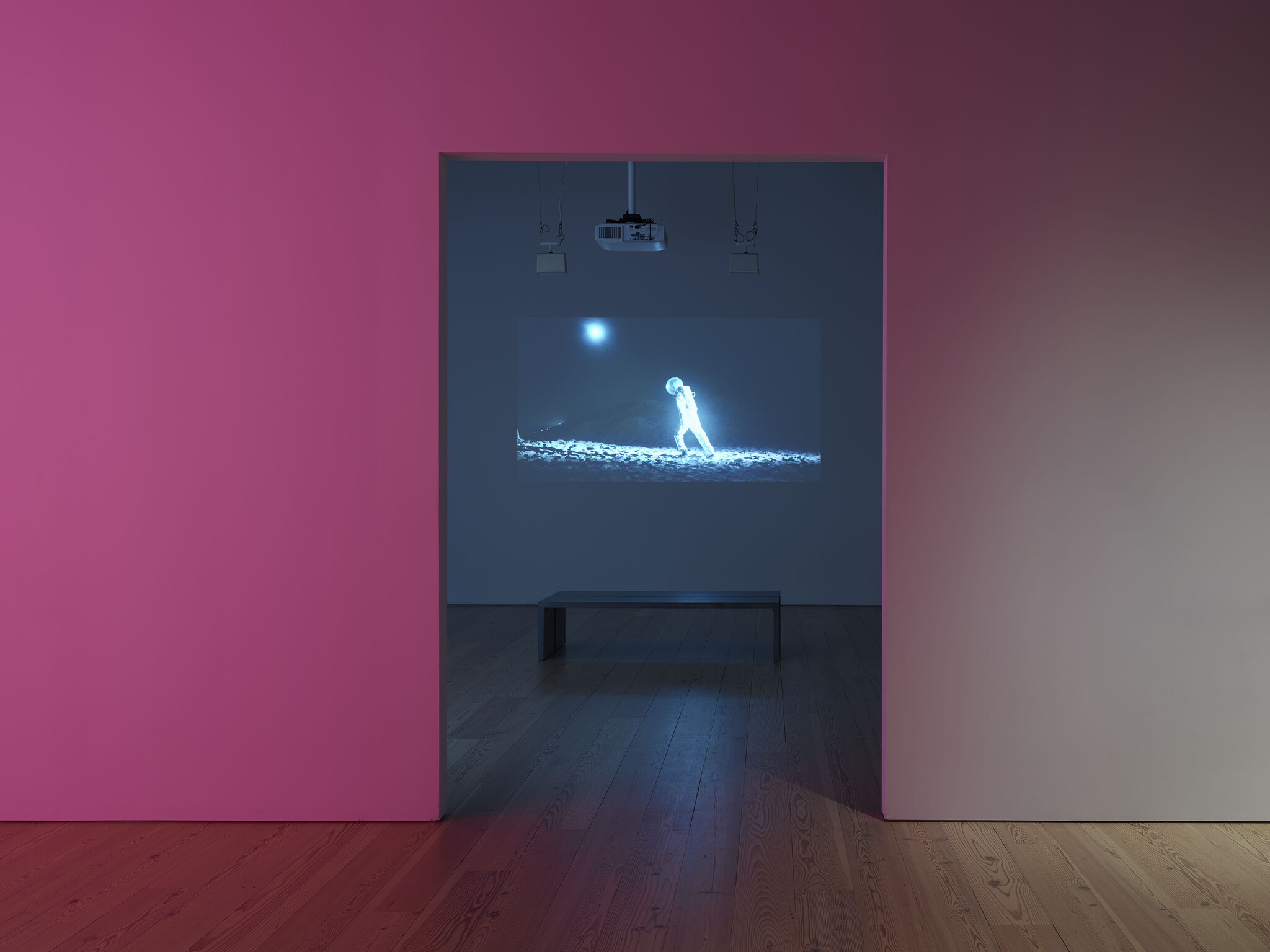 A dark gallery room shows a projected blue astronaut walking on a lunar surface, viewed through a pink doorway.