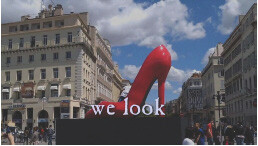 A giant red high-heeled shoe sculpture towers over a city square with the words "we look" visible.