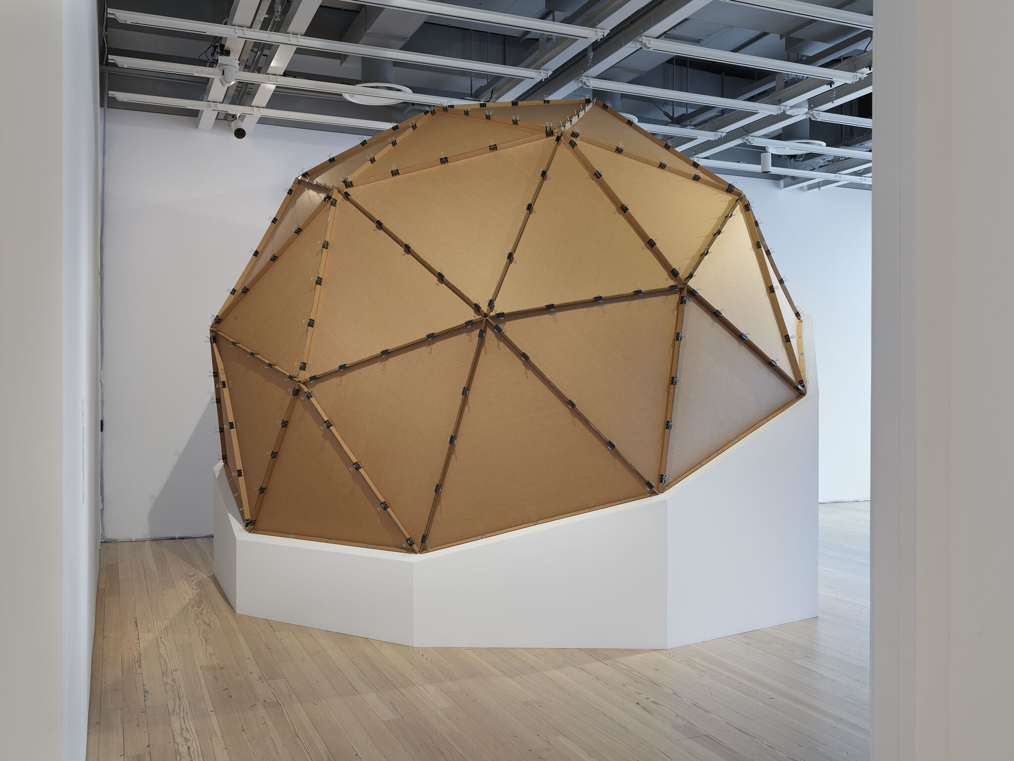 A large geodesic cardboard dome sculpture sits on a white raised platform in a gallery.