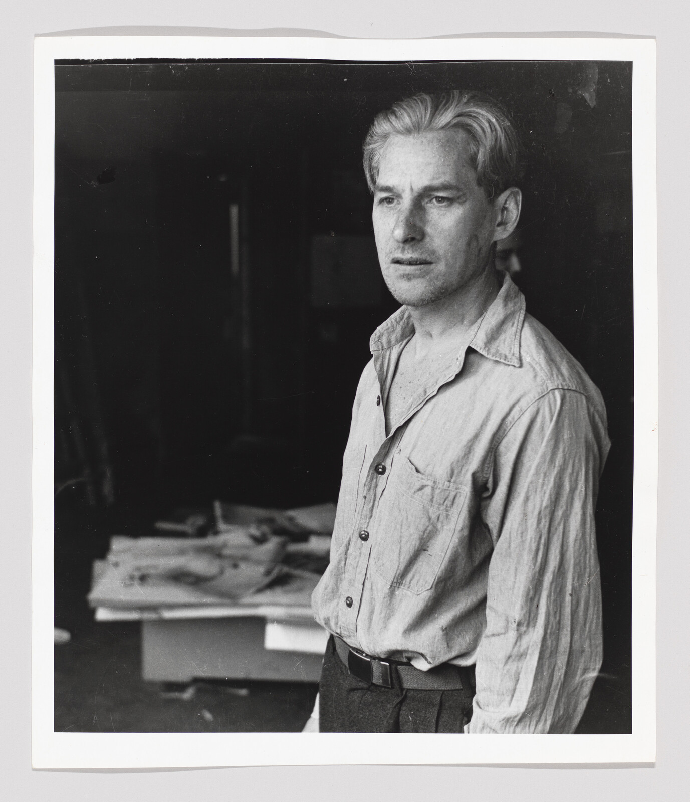 A man in a wrinkled button-up shirt stands in a dim workshop, looking thoughtfully into the distance.