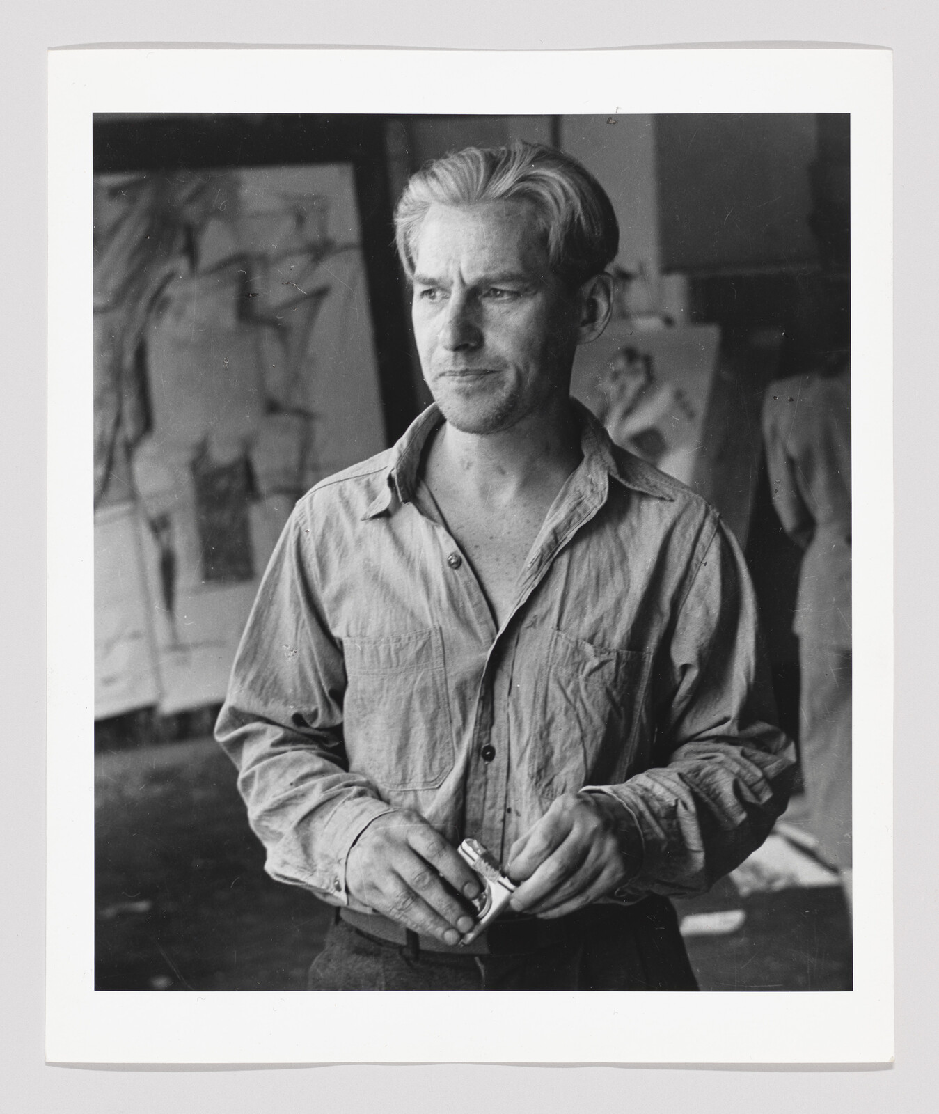 A man in a paint-stained shirt stands in a studio holding a tube of paint.