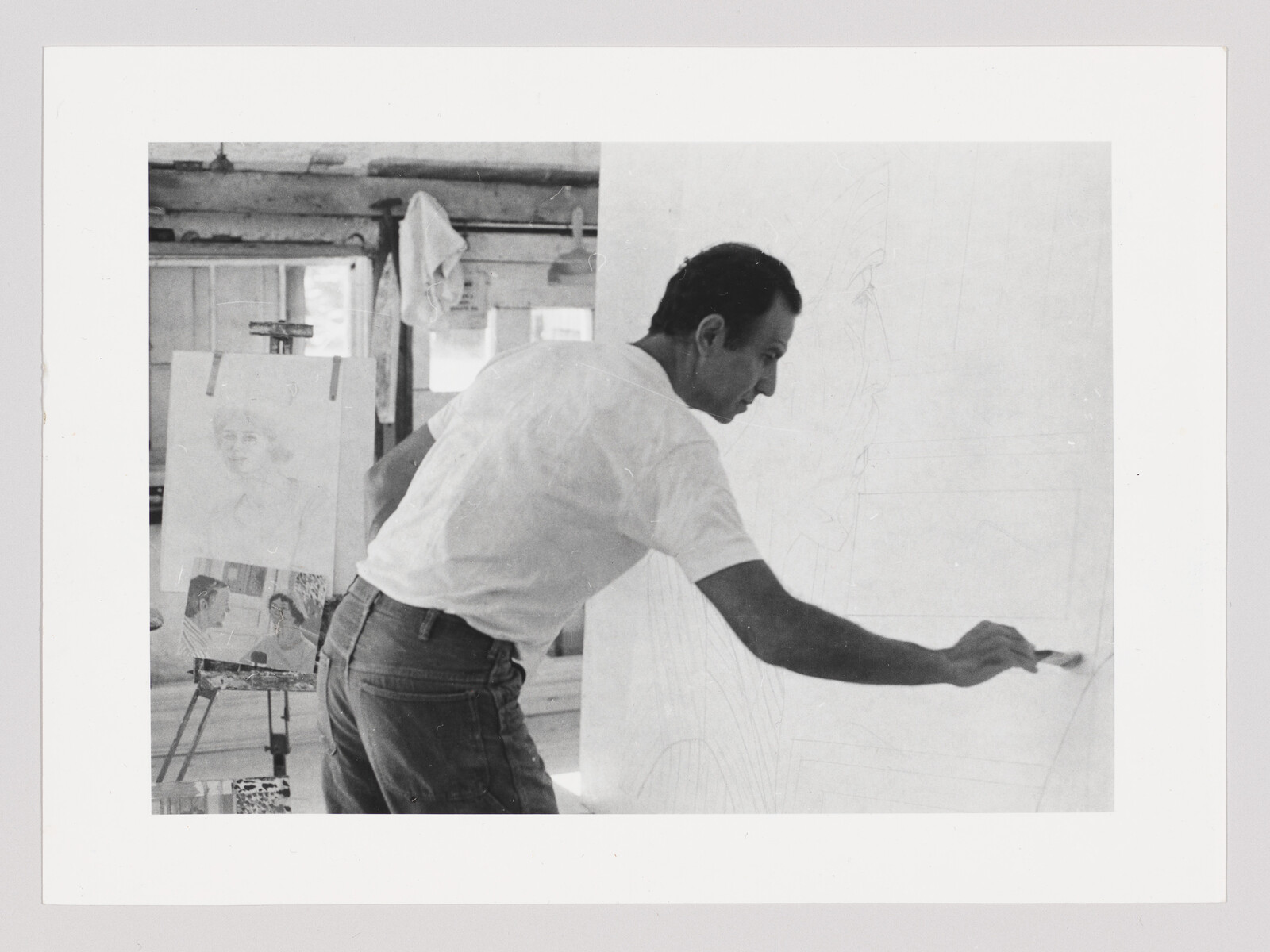 Man painting on a large canvas with a brush in a studio, easel sketches nearby.