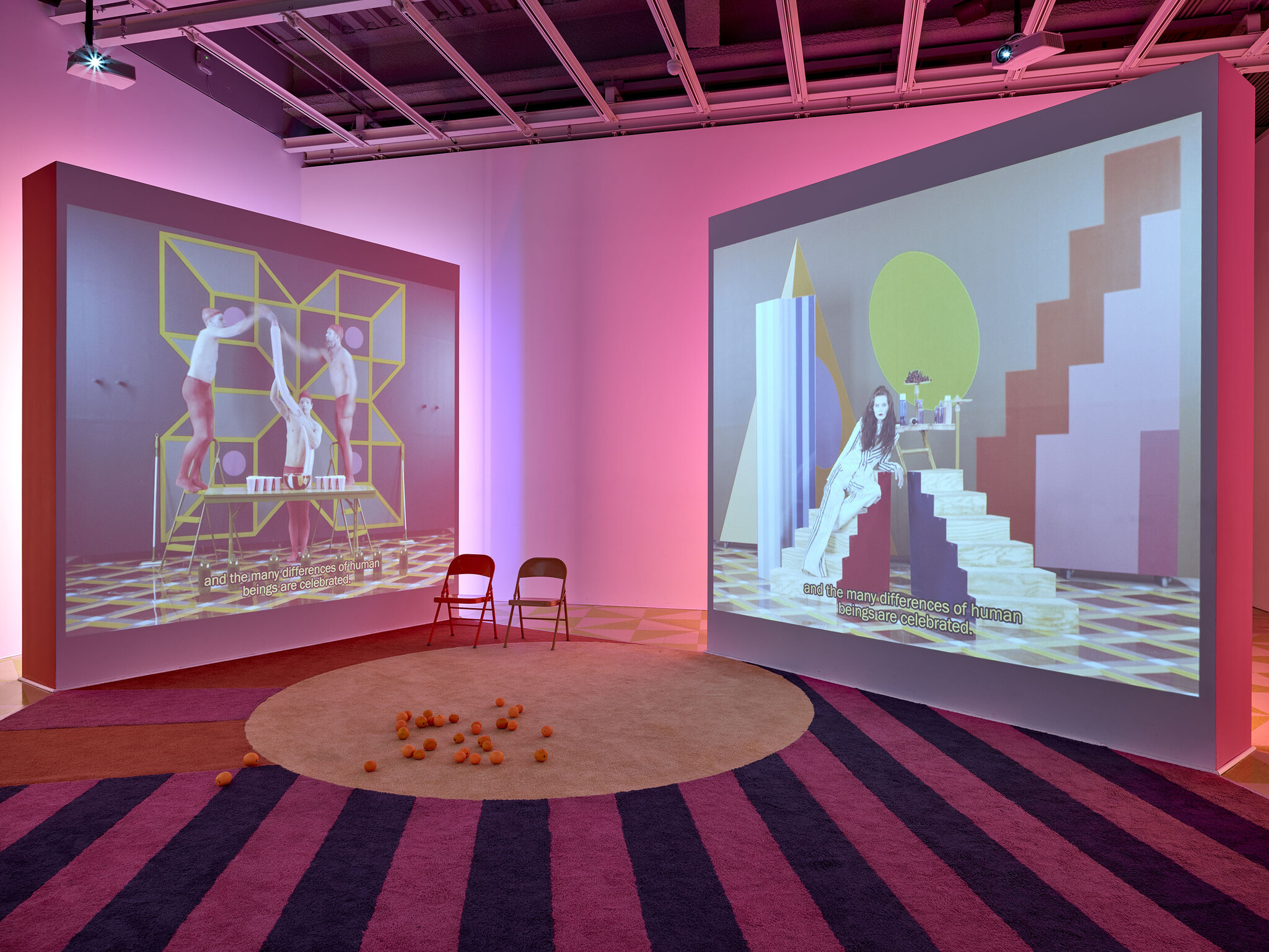 Two large video screens show colorful scenes with people and the subtitle “and the many differences of human beings are celebrated.”