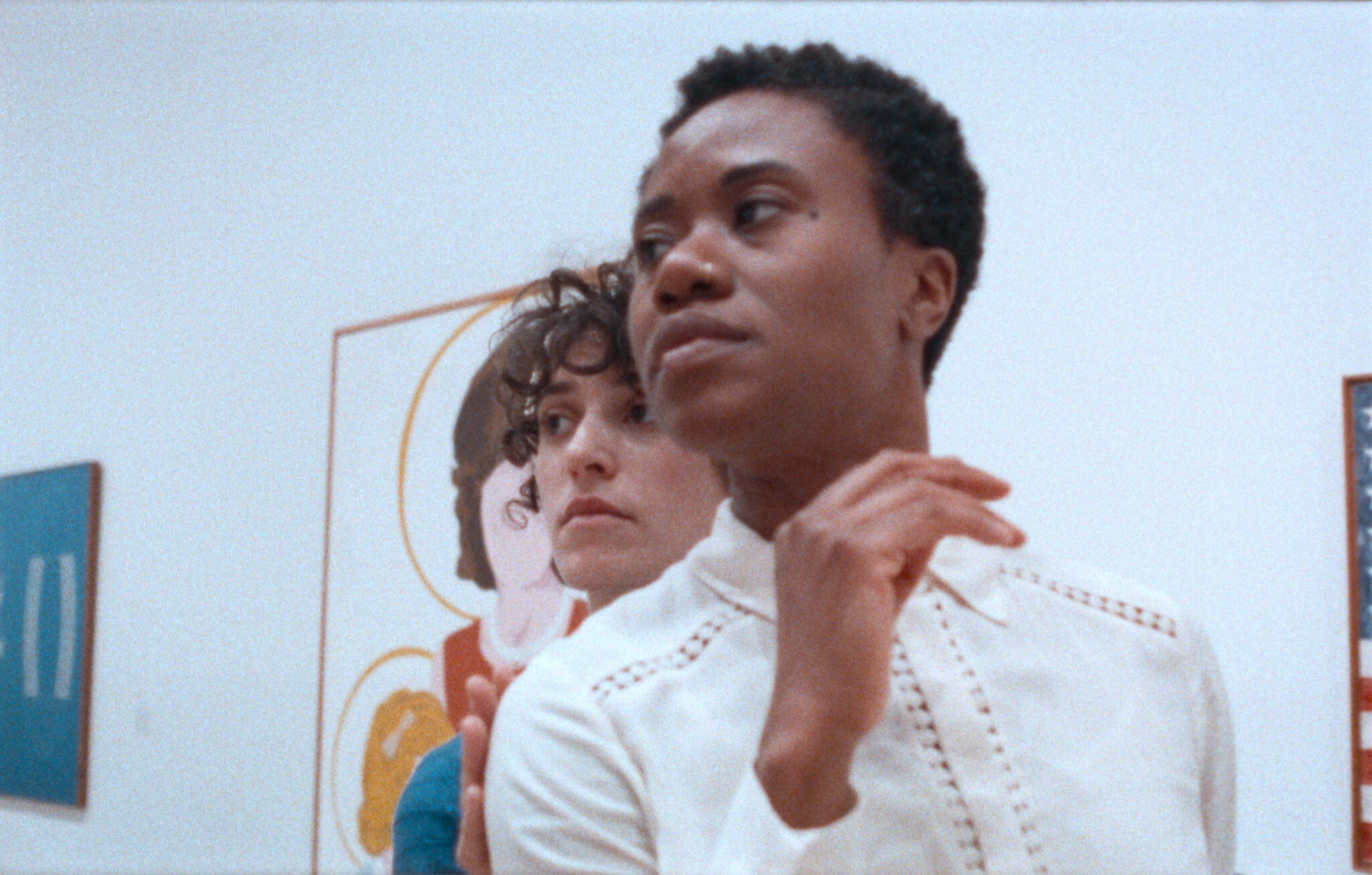Two people stand close together in an art gallery, looking in the same direction with serious expressions.