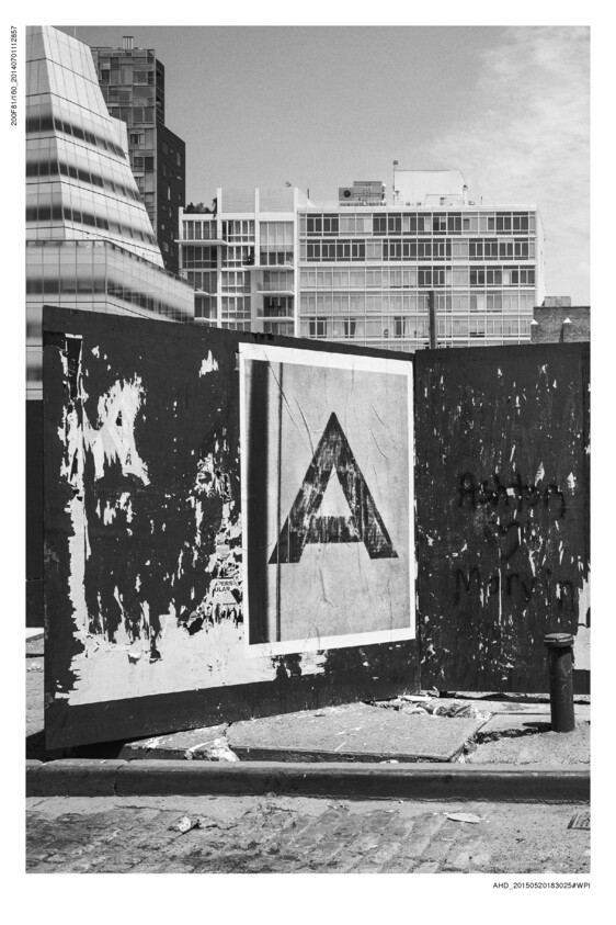 A black and white photograph capturing an urban scene with a large poster featuring a bold triangle design on a wall. The wall shows signs of wear and tear with peeling layers and graffiti. In the background, modern and mid-century style buildings rise against a clear sky. The foreground includes a curb and a paved street, contributing to the gritty aesthetic of the scene.
