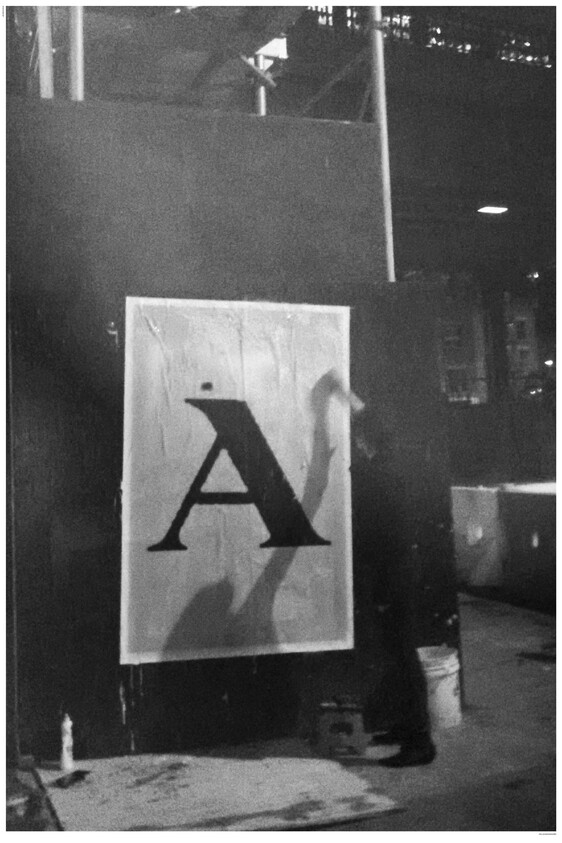 A worker pastes a large printed letter "A" poster onto a dark wall at night.
