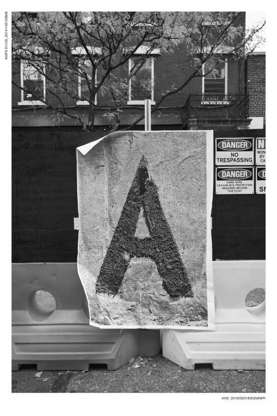 A black and white photograph displaying a large poster with a textured letter 'A' on it, mounted on a barricade. In the background, there's a leafless tree in front of a building with a balcony, and multiple warning signs, including 'DANGER NO TRESPASSING' and 'DANGER HARD HATS, EAR AND EYE PROTECTION REQUIRED BEYOND THIS POINT' are visible on a construction site fence.