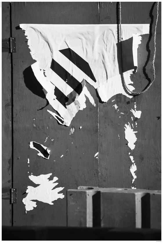 A torn and peeling poster on a wooden surface with a geometric pattern, partially obscured by shadows, in black and white.