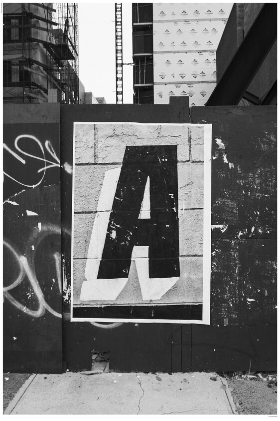 A black and white photograph displaying a large, weathered poster of the letter 'A' affixed to a textured urban wall covered with various layers of peeling paint and graffiti. The scene includes a glimpse of a construction site in the background, with a partially visible building covered in protective sheathing.