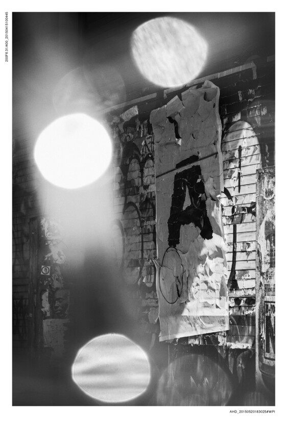A black and white photograph featuring a textured wall covered with layers of torn posters and graffiti. Large, out-of-focus light orbs dominate the foreground, creating a bokeh effect that partially obscures the detailed background.