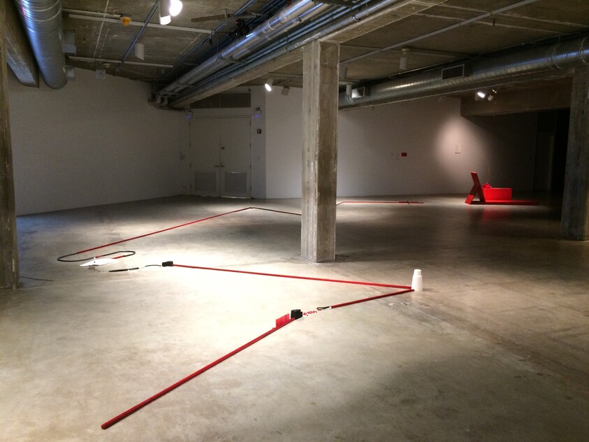 An empty concrete gallery with red tape lines on the floor leading to a red chair sculpture.