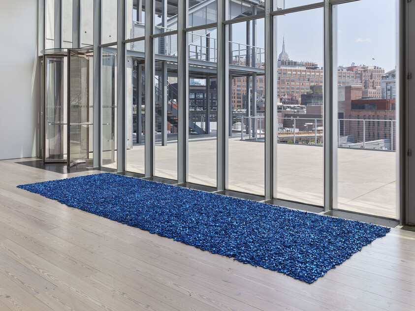 A large rectangular pile of shiny blue material is arranged on a wooden floor near tall glass windows.