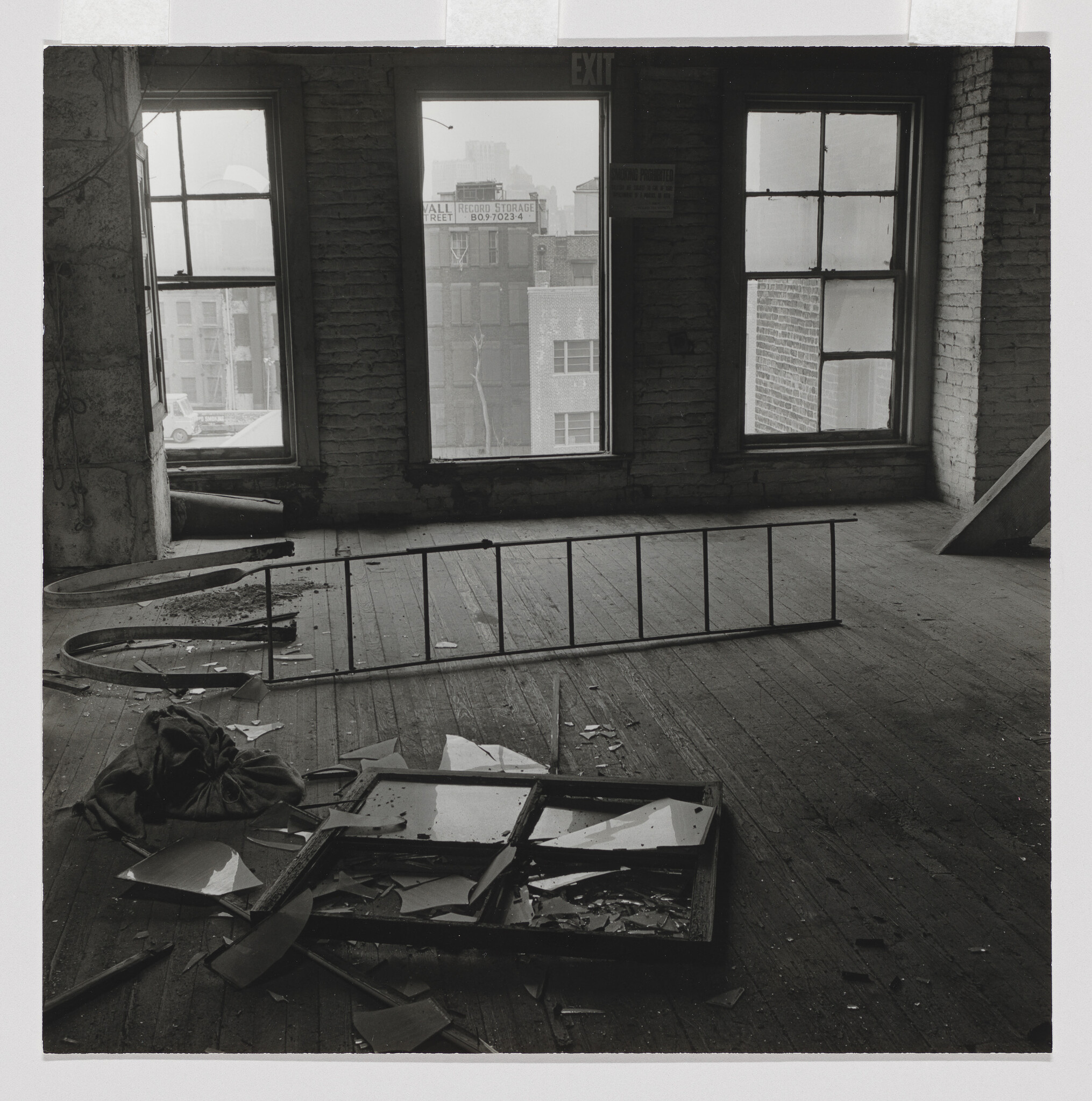 Abandoned room with broken windows, shattered glass on the floor, and a fallen ladder. Two large windows overlook a cityscape.