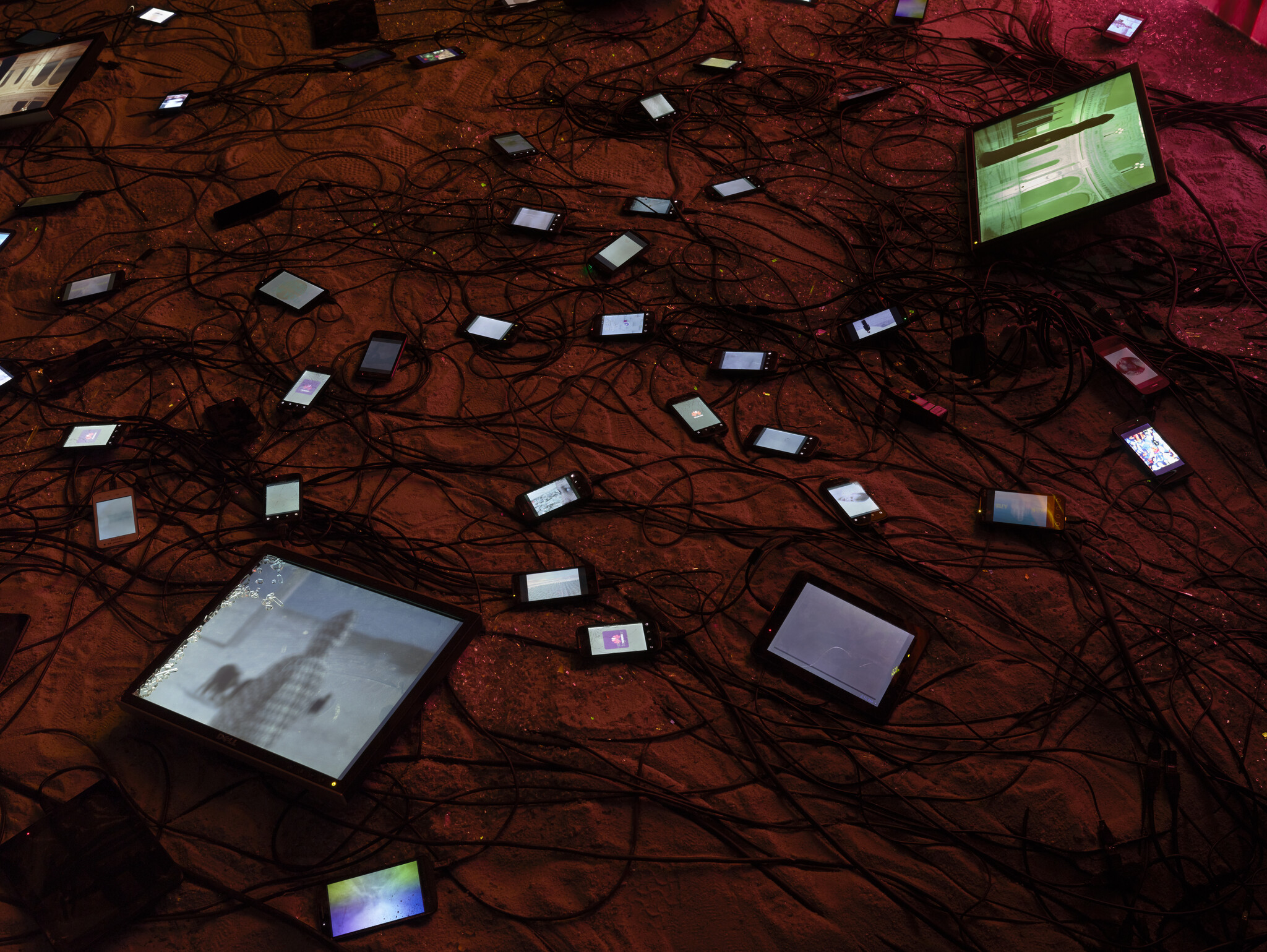 Many smartphones and screens are lying on the ground, connected by tangled black cables, some displaying images.