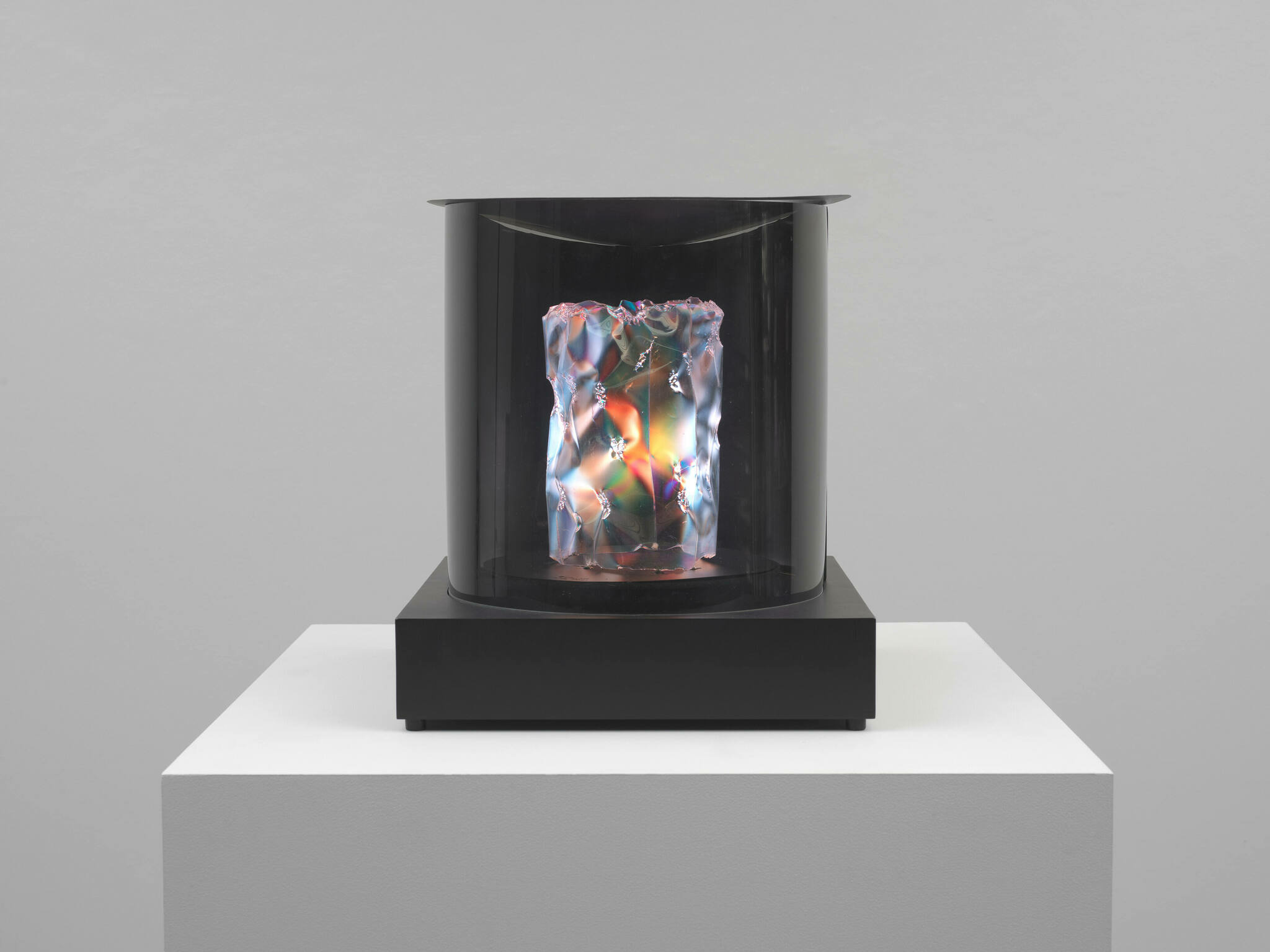 A modern art installation featuring a cylindrical glass case on a black pedestal, with a luminous, multi-colored, abstract sculpture inside. The sculpture emits a bright glow, casting reflections and refractions on the glass surface, set against a plain white gallery wall.