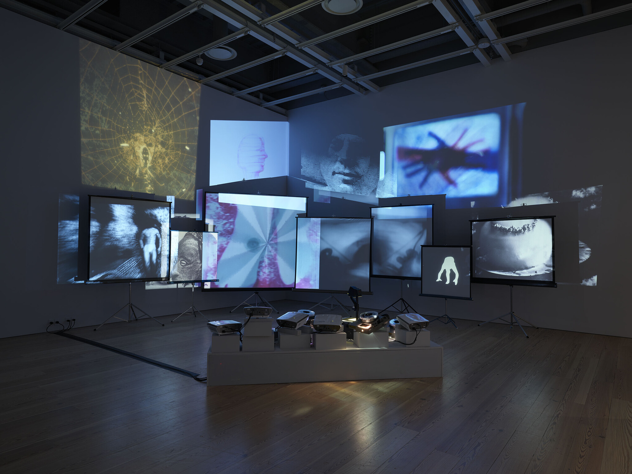 Multiple projectors cast layered abstract and figurative images onto several screens in a dark gallery.
