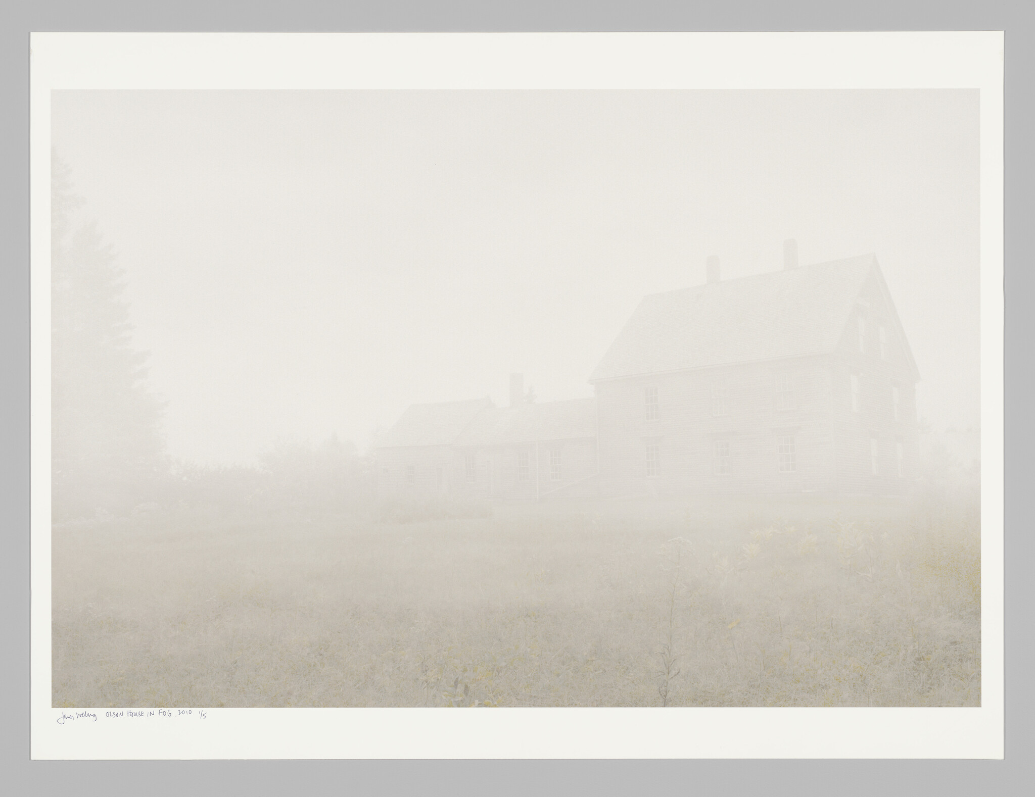 A large farmhouse stands faintly visible through thick morning fog over an empty grassy field.