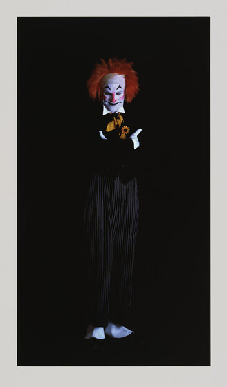 A figure dressed as a clown with exaggerated white makeup, a red nose, and wild red hair stands against a black background. The clown wears a black suit with pinstripes, a yellow bow tie, and white gloves, and is holding a small bouquet of flowers. The expression on the clown's face is a mix of sadness and contemplation.