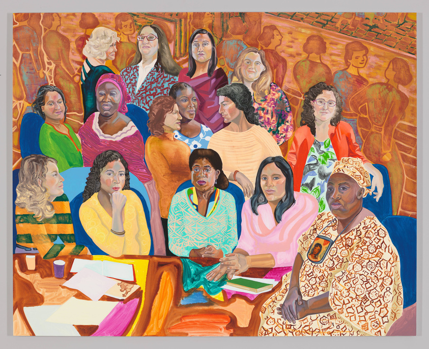A racially diverse group of 15 women in bright business casual outfits and traditional garb sit at and stand around a table. Some look at the viewer and others converse with each other.