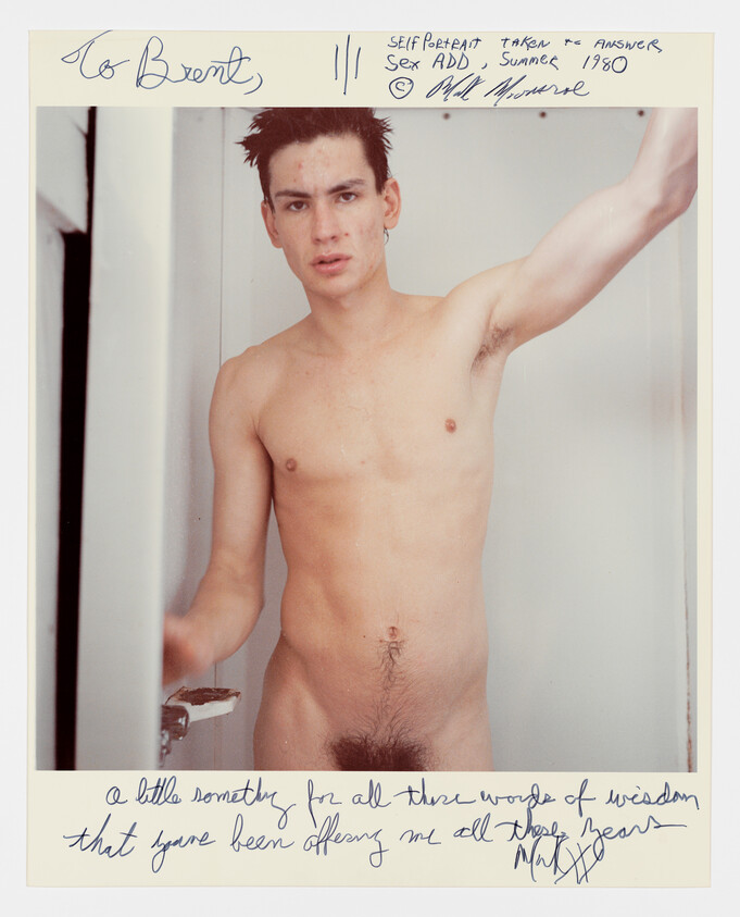 Naked person standing in a doorway looking at the camera with one arm raised.