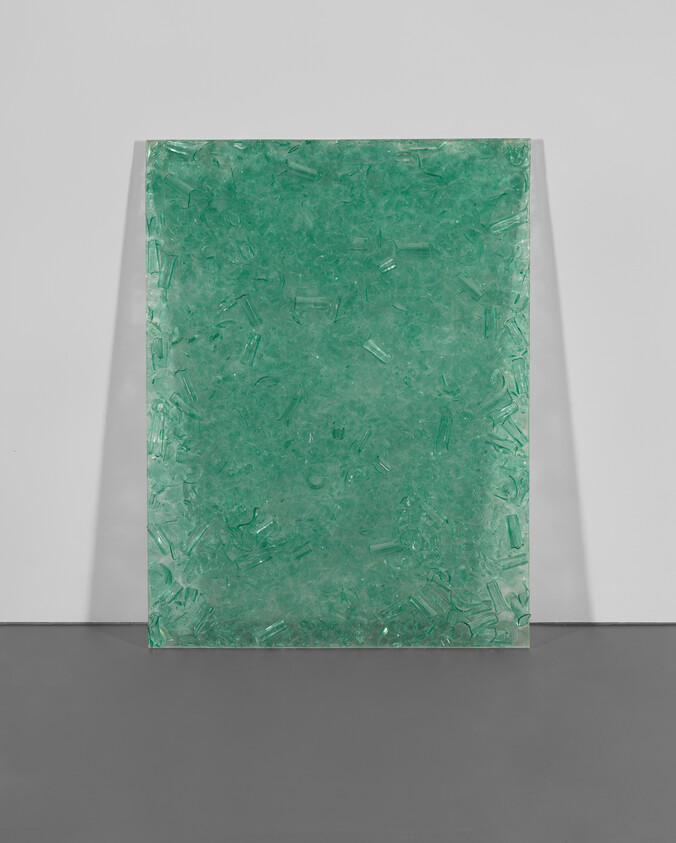 A translucent green panel with embedded objects is displayed upright against a white gallery wall and a grey floor. The panel's texture appears to be rough with various shades of green and internal details that suggest a composition of multiple fragments or materials.