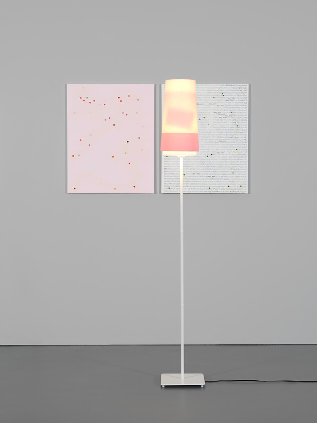 A lit white floor lamp stands in front of two pastel abstract canvases on a gray wall.