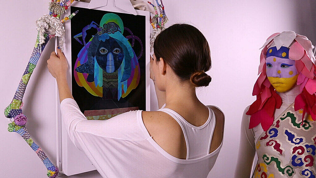 A woman adjusts a colorful framed artwork while a person in a bright, decorative costume stands nearby watching.